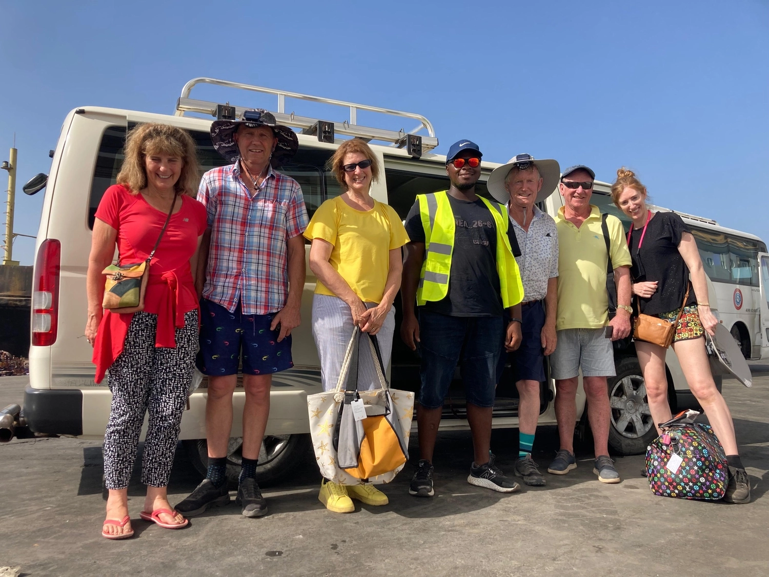 Gambia View Tours - Tourism services - A group of seven diverse tourists and a guide in a high-visibility vest standing together in front of a white tour van on a sunny day.